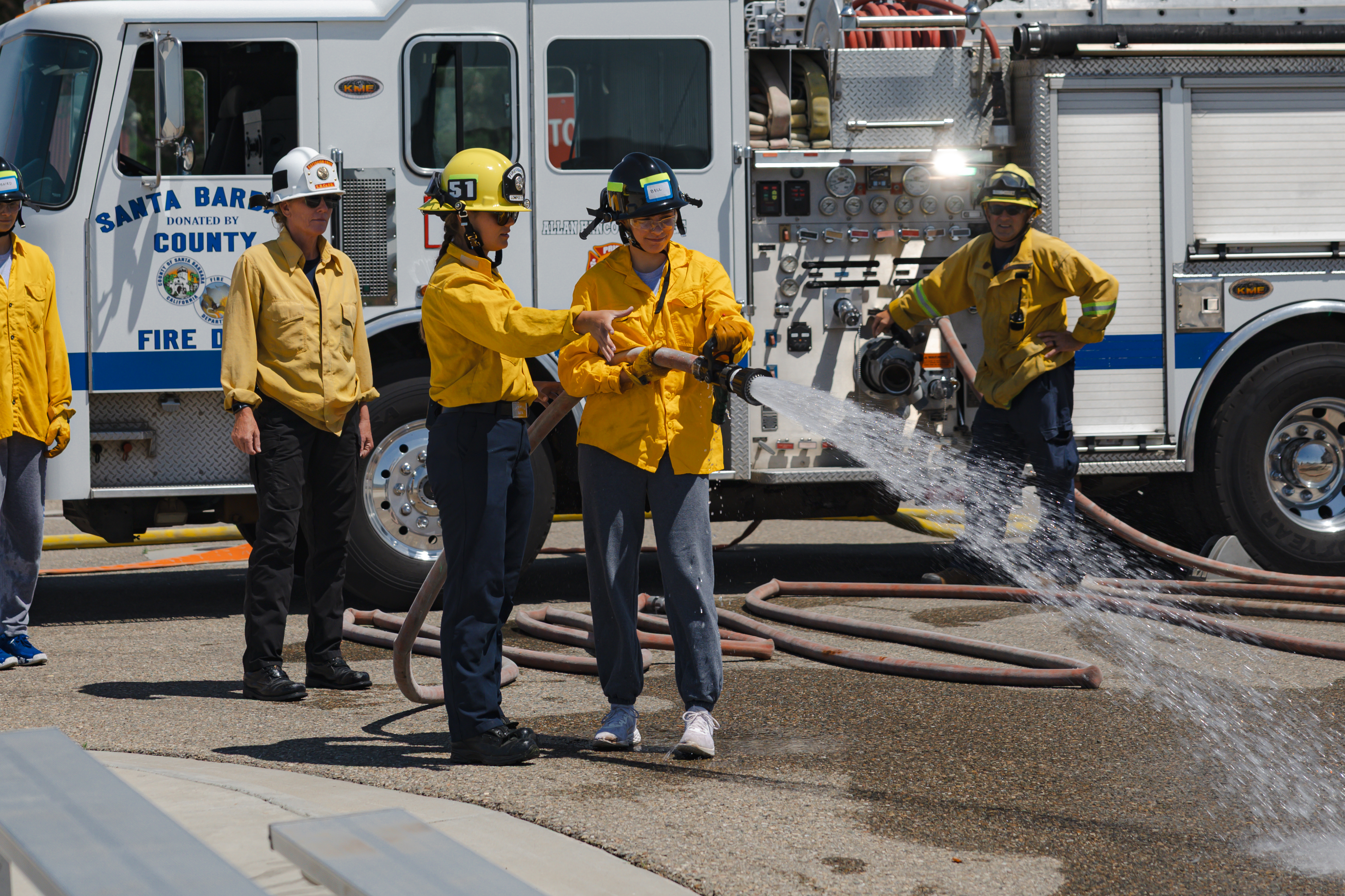 Participants in Allan Hancock College’s Women’s Fire Camp practice operating a fire hose beside a fire engine during a hands-on training exercise. Wearing yellow protective fire shirts and helmets, the participants spray water while instructors and other trainees observe nearby.